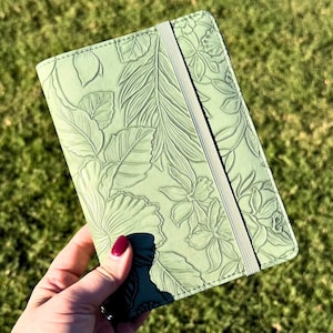 May include: A light green journal with an embossed floral design. The cover features a detailed pattern of leaves and flowers, with an elastic band closure. The journal is held against a backdrop of green grass.