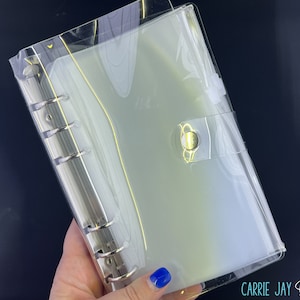 May include: A clear plastic binder with a gold button closure. The binder has six rings and is designed to hold loose leaf paper.