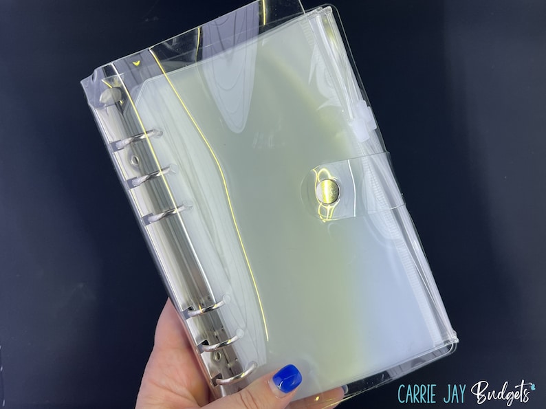 May include: A clear plastic binder with a gold button closure. The binder has six rings and is designed to hold loose leaf paper.
