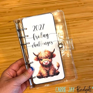 Puede incluir: Una carpeta de plástico transparente con mecanismo de anillas metálicas, que incluye una hoja blanca con el texto 2027 friday challenges y una ilustración gráfica de un ternero Highland marrón con pelaje desgreñado.