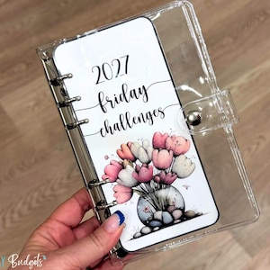 May include: A clear plastic six-ring binder featuring a white insert with the text 2027 friday challenges and a colorful illustration of pink and white tulips in a glass vase filled with stones, secured by a silver metal snap button closure.