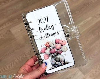 Può includere: Un raccoglitore trasparente a sei anelli con un inserto bianco recante il testo 2027 friday challenges e un'illustrazione colorata di tulipani rosa e bianchi in un vaso di vetro riempito di pietre, chiuso da un bottone a pressione in metallo.
