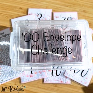 May include: A clear plastic container with a lid, filled with envelopes. The container has a black label that reads "100 Envelope Challenge". The container is surrounded by envelopes with numbers written on them.