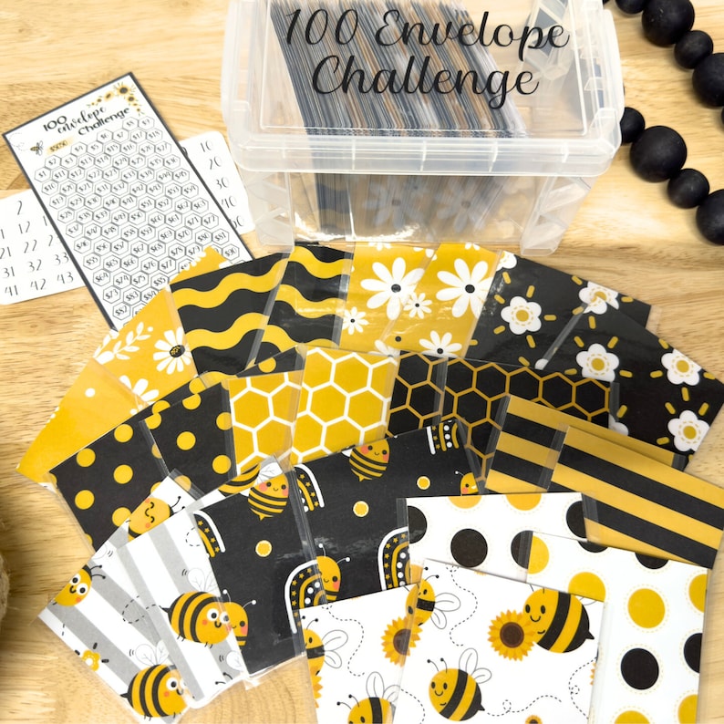 May include: A set of 100 envelopes with various bee-themed patterns, including black and yellow polka dots, honeycomb, stripes, and floral designs. The envelopes are arranged in a grid pattern on a wooden surface. A clear plastic container with the text "100 Envelope Challenge" is in the background.