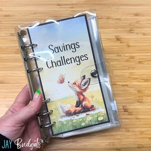 May include: A clear plastic binder with a colorful illustration of a fox and a butterfly. The binder has the words "Savings Challenges" printed on the front.