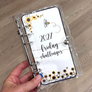 Puede incluir: Una carpeta transparente de seis anillas con una hoja blanca ilustrada con girasoles amarillos, una pequeña abeja y el texto 2027 friday challenges. La carpeta cuenta con un cierre de presión plateado y anillas metálicas.