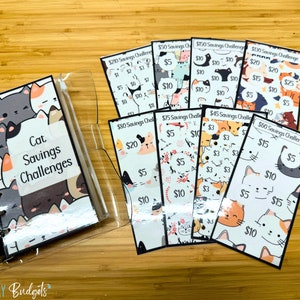 May include: A clear plastic binder with a cat-themed cover that says "Cat Savings Challenges". The binder contains several colorful cards with different amounts of money on them, such as $5, $10, $15, $20, and $25. Each card has a cute cat illustration.