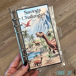 May include: Clear plastic binder with a dinosaur-themed cover. The cover features a colorful illustration of various dinosaurs in a prehistoric landscape, with the text "Savings Challenges" at the top. The binder has a silver metal ring mechanism.