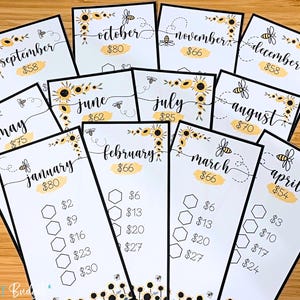 May include: A collection of white cards with black borders, each featuring a month's name in cursive, a dollar amount, and bee and sunflower illustrations. The cards are arranged on a wooden surface.