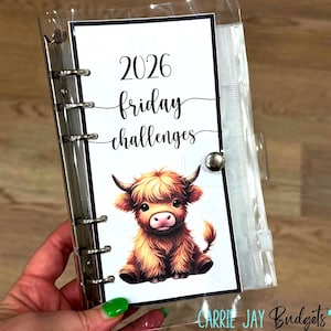 May include: A clear binder with a white insert featuring the text "2026 friday challenges" and a watercolor illustration of a Highland cow. The binder is held by a hand. The text "Carrie Jay Budgets" is at the bottom.