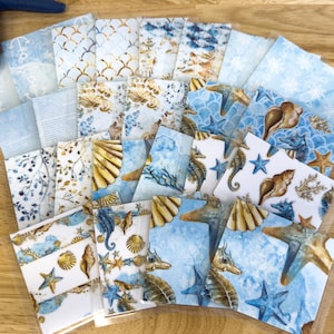 May include: A set of 20 decorative paper sheets featuring a nautical theme. The sheets are printed with various designs including seashells, starfish, seahorses, and ocean waves in shades of blue, white, and gold.
