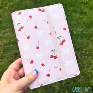 May include: A pink notebook cover with a pattern of red cherries and white bows. The cover has an elastic band closure and is held by a hand. The text "CARRIE JAY BUDGETS" is visible at the bottom right.