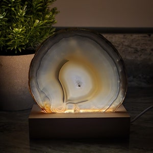 Op de afbeelding: Een witte agaat geode lamp met een houten voet. De geode wordt van binnenuit verlicht, waardoor een warme gloed ontstaat.