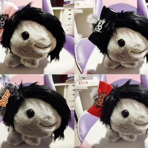 May include: Four plush toys with black hair and different colored bows. The bows have different designs, including a pink bow with a black and white design, a black bow with a white design, a red bow with a black and white design, and a gray bow with the word "Riot" on it.