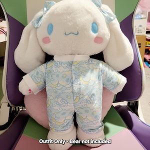 May include: Blue and white pyjamas with a cloud and moon pattern for a plush toy. The outfit is for a stuffed animal that is not included. "Outfit Only - Bear not included" is written on the pink surface below the toy.
