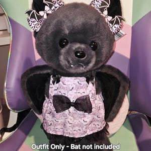 May include: A plush bat toy wearing a pink and black outfit. The outfit includes a patterned top with a black bow and black lace trim. The bat has black ears with bat-shaped bows and the text "Outfit Only - Bat not included".