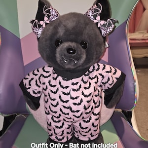 May include: A plush bat outfit in pink with black bat silhouettes. The outfit includes a short-sleeved top and shorts. The bat's ears are adorned with black and pink bows. The text "Outfit Only - Bat not included" is at the bottom.