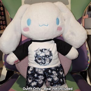 May include: A white plush toy wearing a black and white pyjama set with a ghost print. The pyjama top is white with a black and white ghost print. The pyjama trousers are black with a white ghost print. The text "Outfit Only - Bear not included" is at the bottom of the image.