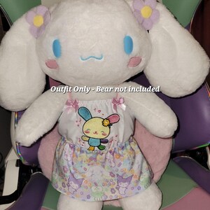 May include: A white plush toy wearing a pink and white dress with a colorful embroidered character on the front. The dress has a floral print skirt. The text "Outfit Only - Bear not included" is visible on the image.