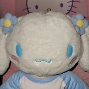 May include: A plush white character with large ears, blue eyes, and a stitched blue mouth. The character has two blue and yellow flower accents and is wearing a blue and white shirt. The background is pink.