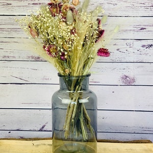May include: A grey glass vase holding a dried flower bouquet. The arrangement features white, burgundy, and beige flowers and grasses. The vase is a smoky grey colour and rests on a wooden surface, set against a white wood-panelled backdrop.