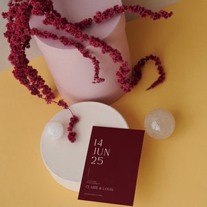 May include: A burgundy save the date card with white text that reads "14 JUN 25" and "Save the date for the wedding of CLAIRE & LOUIS". The card is on a white round surface with a small white sphere and a clear crystal sphere. The background is a yellow surface.