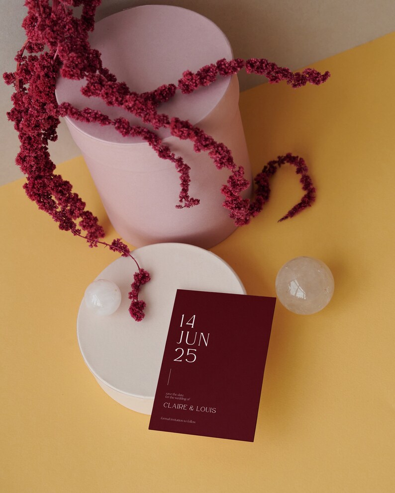 May include: A burgundy save the date card with white text that reads "14 JUN 25" and "Save the date for the wedding of CLAIRE & LOUIS". The card is on a white round surface with a small white sphere and a clear crystal sphere. The background is a yellow surface.