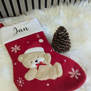 May include: Red Christmas stocking with a white cuff embroidered with the name "Jan". The stocking features a beige teddy bear wearing a Santa hat and a red bow. White snowflakes and dots decorate the stocking. A pine cone sits next to the stocking.