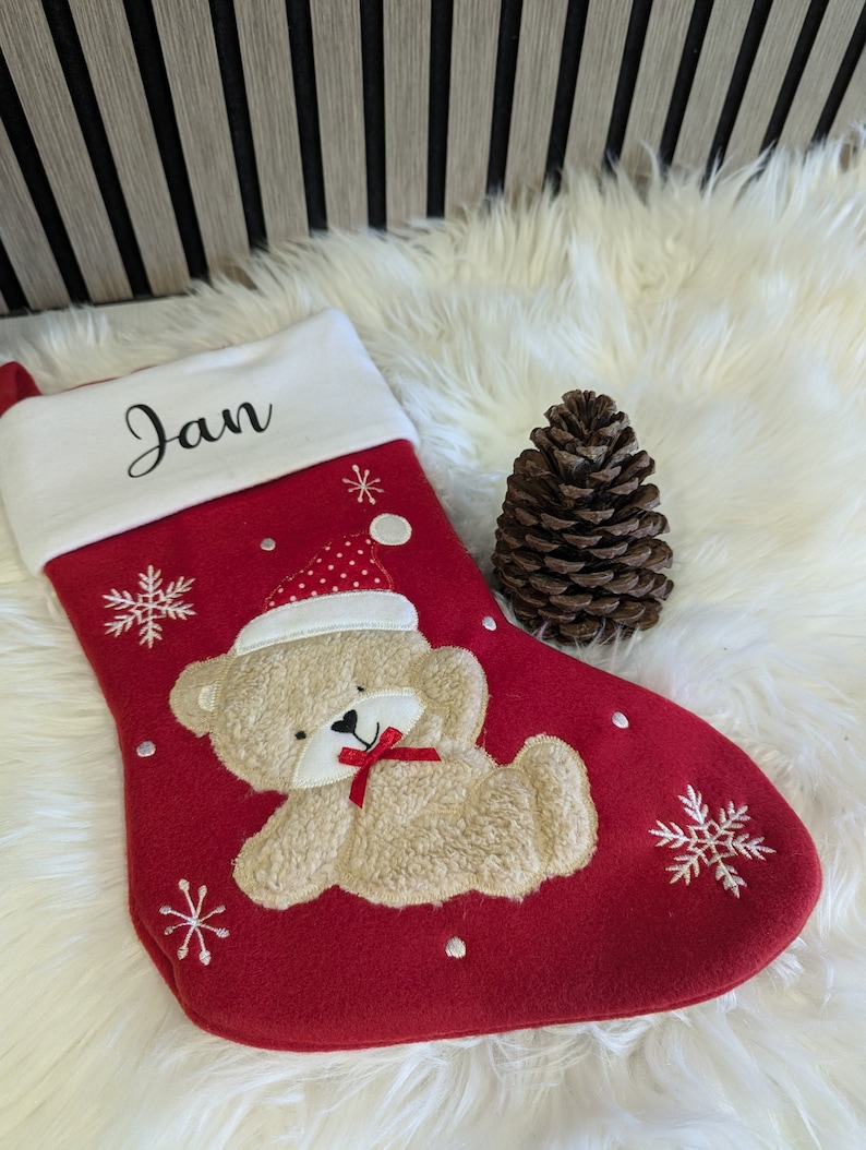 May include: Red Christmas stocking with a white cuff embroidered with the name "Jan". The stocking features a beige teddy bear wearing a Santa hat and a red bow. White snowflakes and dots decorate the stocking. A pine cone sits next to the stocking.