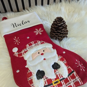 May include: A festive red Christmas stocking with a Santa Claus design and the name "Niclas" embroidered on the white cuff. The stocking is decorated with snowflakes and a black belt with a gold buckle. A brown pine cone is also present.