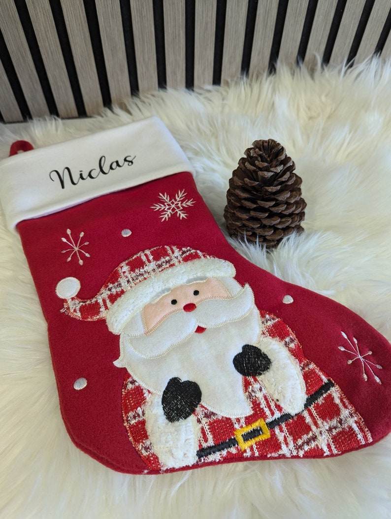 May include: A festive red Christmas stocking with a Santa Claus design and the name "Niclas" embroidered on the white cuff. The stocking is decorated with snowflakes and a black belt with a gold buckle. A brown pine cone is also present.