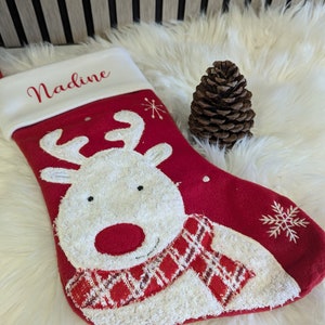 May include: A festive red Christmas stocking with a white cuff, featuring a reindeer design and the name "Nadine". The reindeer has a white face, red nose, and a red and white plaid scarf. A brown pine cone is placed beside the stocking.