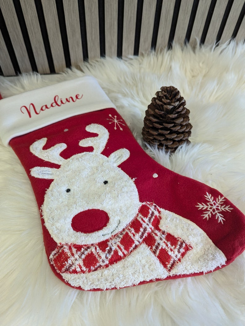 May include: A festive red Christmas stocking with a white cuff, featuring a reindeer design and the name "Nadine". The reindeer has a white face, red nose, and a red and white plaid scarf. A brown pine cone is placed beside the stocking.