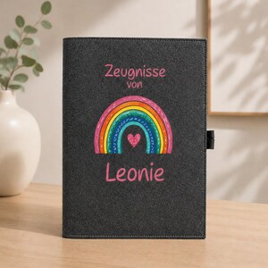 Könnte beinhalten: Eine dunkelgraue Filzmappe mit der Aufschrift Zeugnisse von Leonie in rosa Buchstaben. Zentriert unter dem Text befindet sich eine bunte Regenbogen-Grafik mit einem kleinen rosa Herz an der Basis. Die Mappe hat eine schwarze Stiftschlaufe.