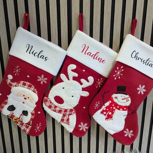 May include: Three red Christmas stockings with white cuffs, each bearing a name: Niclas, Nadine, and Christian. The stockings are decorated with festive designs: Santa Claus, a reindeer, and a snowman. They hang against a striped backdrop.