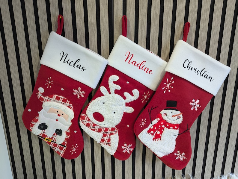 May include: Three red Christmas stockings with white cuffs, each bearing a name: Niclas, Nadine, and Christian. The stockings are decorated with festive designs: Santa Claus, a reindeer, and a snowman. They hang against a striped backdrop.