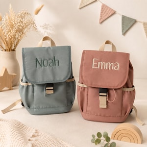 Könnte beinhalten: Zwei kleine Rucksäcke, einer in Staubblau und einer in Rosa, mit den Namen "Noah" und "Emma" auf den vorderen Klappen gestickt. Jeder hat einen beigen Griff und Besatz, eine schwarze Schnalle und Seitentaschen. Ein bezauberndes Accessoire für Kinder.