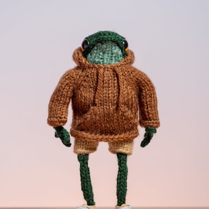 Könnte beinhalten: Ein grüner und brauner gestrickter Frosch, der ein braunes Hoodie, braune Shorts und gelbe Turnschuhe trägt.