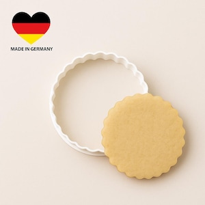 Könnte beinhalten: Ein weißer Ausstecher mit gewelltem Rand neben einem runden, hellbraunen Keks. Das Bild enthält ein herzförmiges Grafik in den Farben der deutschen Flagge mit dem Text "MADE IN GERMANY".
