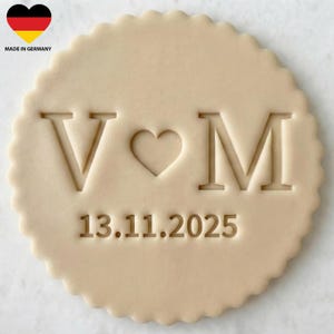 May include: A beige, round cookie cutter with a scalloped edge. The design includes the letters "V" and "M" separated by a heart shape, and the date "13.11.2025" below. The upper left corner shows a German flag.