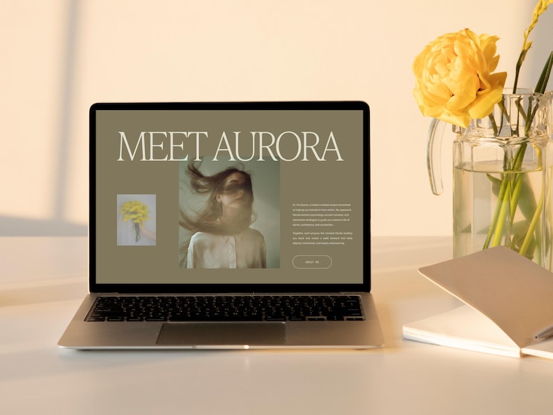 May include: A laptop displaying a website with the text "MEET AURORA" and a photo of a person with flowing hair. A bouquet of yellow flowers in a glass vase sits to the right, with a notebook and pen nearby. The scene is bathed in warm light.