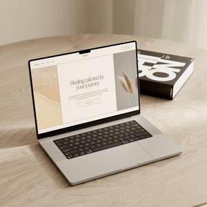 May include: A silver laptop displaying a website with the text "Healing tailored to your journey" on a light wood table. A black book with white text is next to the laptop.