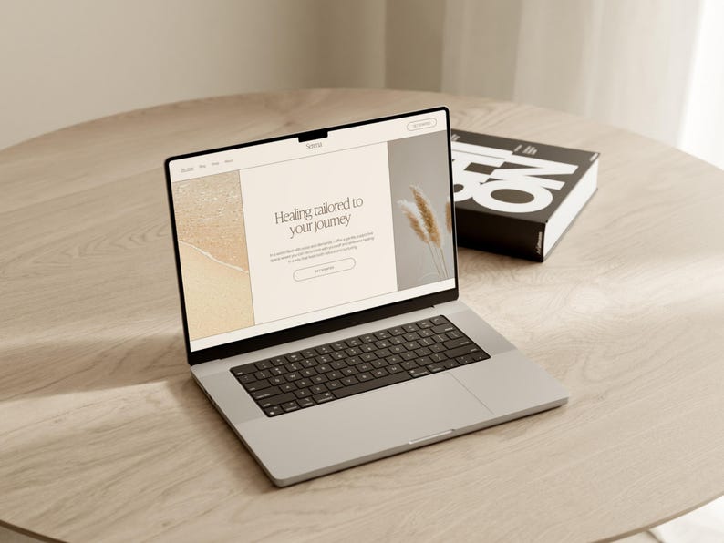 May include: A silver laptop displaying a website with the text "Healing tailored to your journey" on a light wood table. A black book with white text is next to the laptop.