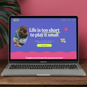 May include: A silver MacBook Pro laptop showing a website with the text "Life is too short to play it small." The website has a blue background with pink and yellow accents. The laptop is on a wooden surface.