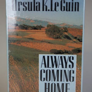 May include: A book cover with a blue background and the title "ALWAYS COMING HOME" in black text. The author's name, "Ursula K. Le Guin", is printed in black text at the top of the cover. The cover features a photograph of a grassy field with rolling hills in the background.