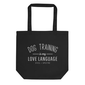以下が含まれることがあります： 「DOG TRAINING is my LOVE LANGUAGE」と「STEEL + SPECTRE」の白い文字が入った黒いトートバッグ。バッグには2つのハンドルがあり、丈夫な素材で作られています。物を運ぶのに適しています。