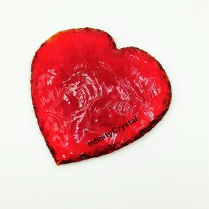 May include: A red heart-shaped piece of glass with a swirling pattern. The glass is translucent and has a slight orange hue. The text "InfinityCrystal" is printed on the bottom of the heart.