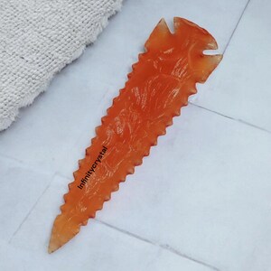 May include: Orange crystal point with a serrated edge and a textured surface. The crystal is shaped like a spearhead and has a pointed tip. The text "infinitycrystal" is written on the crystal.