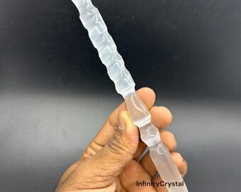 May include: A clear, elongated crystal wand with a ridged, cylindrical center and a pointed tip. The wand is held against a dark background. The text "InfinityCrystal" is visible on the image.