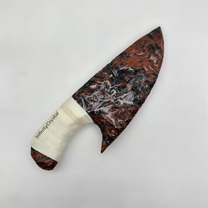 May include: A decorative knife with a textured, reddish-brown blade and a white, wrapped handle. The blade has black and white marbling. The text "InfinityCrystal" is printed on the handle.